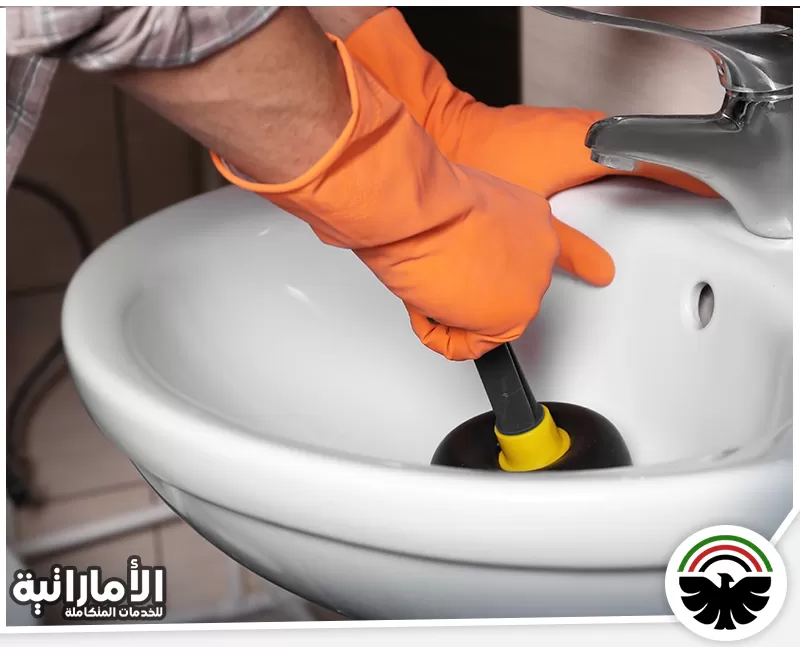 طريقة فتح المجاري الحمام 0582600901 المؤسسة الإماراتية فني يستخدم مكبس اليد لتسليك مجاري الحوض، مرتديًا قفازات لحماية يديه أثناء العمل.