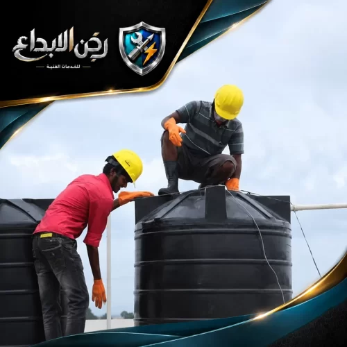 عاملان على سطح مبنى يقومان بتركيب أو إصلاح خزانات مياه بلاستيكية سوداء، مرتديين خوذات صفراء