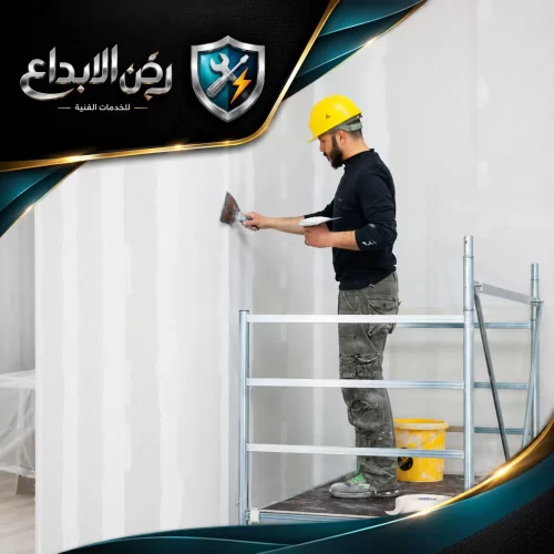 عامل يرتدي خوذة صفراء يقف على سقالة معدنية لمعالجة فواصل الجدران بدقة ضمن أعمال معجون ولياسة الجدران